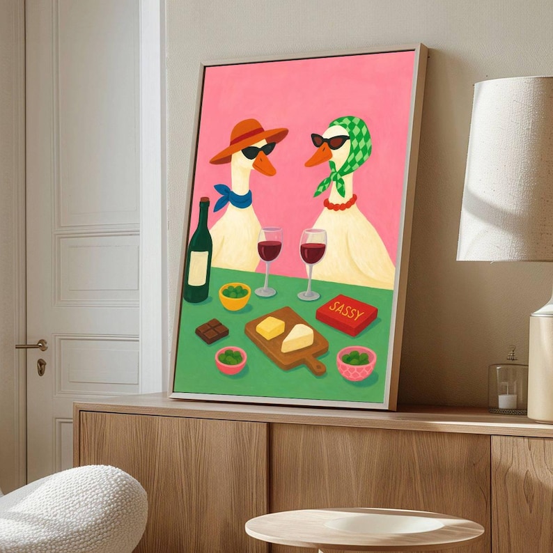May include: A framed art print featuring two geese wearing sunglasses, hats, and scarves, enjoying wine and snacks. The artwork has a pink background and a green table with the word "SASSY" on a red box.