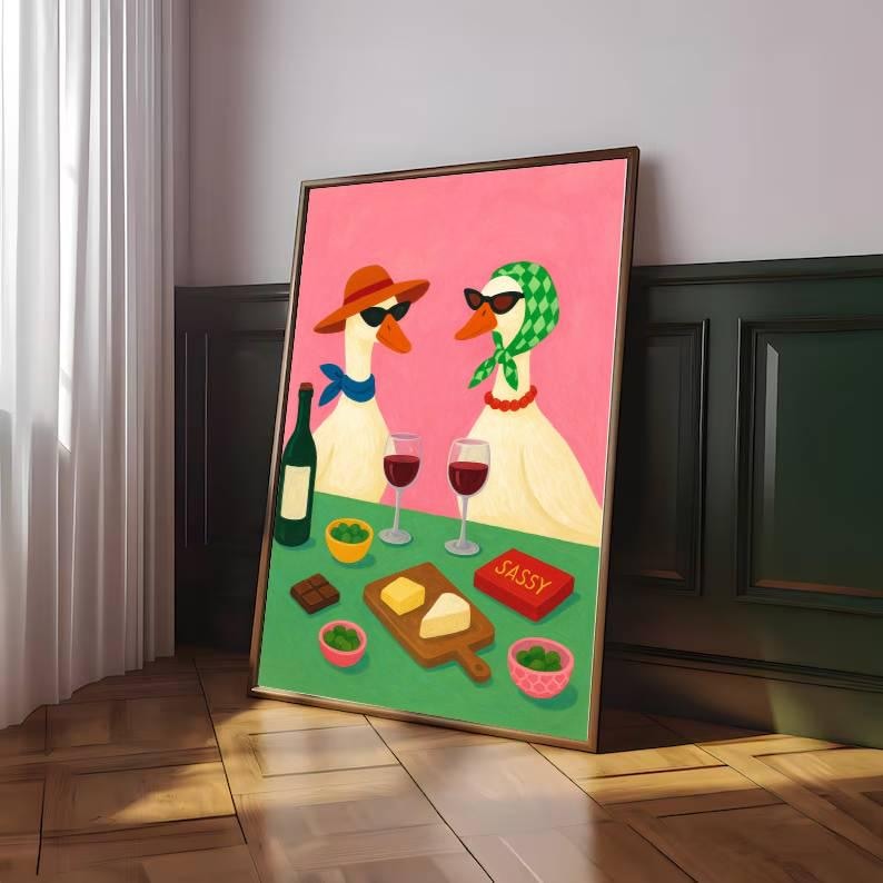 May include: A framed art print featuring two geese wearing sunglasses and accessories, enjoying wine and snacks. The geese are set against a pink background, with a green table holding wine glasses, cheese, and the word "SASSY" on a box.
