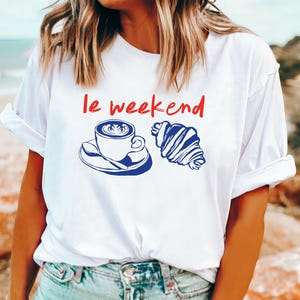 May include: White t-shirt with the phrase "le weekend" in red. A blue illustration of a coffee cup and croissant is printed below the text. The shirt has rolled-up sleeves.