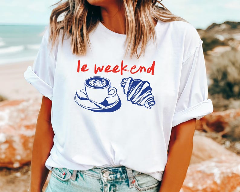 May include: White t-shirt with the phrase "le weekend" in red. A blue illustration of a coffee cup and croissant is printed below the text. The shirt has rolled-up sleeves.
