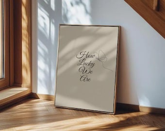 May include: A framed art print with the words "How Lucky We Are" in elegant script, accompanied by a delicate butterfly illustration. The artwork has a neutral color palette and is displayed on a wooden floor.