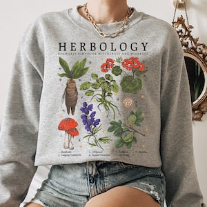 May include: A gray sweatshirt with a graphic print of various plants and the text "HERBOLOGY HOGWARTS SCHOOL OF WITCHCRAFT AND WIZARDRY". The graphic includes a mandrake, toadstools, gillyweed, fanged geranium, a fig, and aconite.