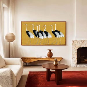 May include: A framed artwork featuring six stylized cranes in black, white, and gray against a golden yellow background. The artwork is displayed above a wooden console table in a living room setting. The room also includes a red rug, a white sofa, and a round coffee table.