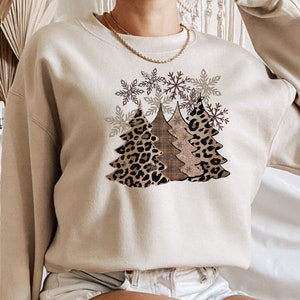 May include: A beige sweatshirt with a design of three Christmas trees. The trees are made of leopard print fabric, brown plaid fabric, and a glittery gold fabric. The trees are surrounded by snowflakes.