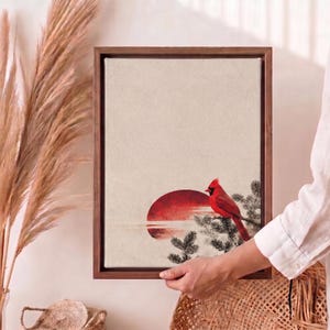 May include: A framed canvas art print featuring a red cardinal perched on a branch, set against a red sun and a neutral background. The artwork is encased in a dark wooden frame, creating a serene and natural aesthetic.
