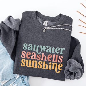 May include: A dark gray sweatshirt with the words "saltwater seashells sunshine" in teal, coral, and yellow. A silver necklace with a pendant is on the sweatshirt. The sweatshirt is folded on top of a pair of blue denim shorts.