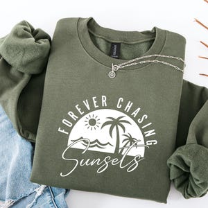 May include: A green sweatshirt with the text "FOREVER CHASING SUNSETS" in white. The graphic includes a sun, palm trees, and waves.