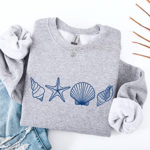 May include: A gray sweatshirt with a crew neck and rolled-up sleeves. The sweatshirt features a blue seashell design across the chest, including a starfish and various seashells. A silver necklace is draped across the top.