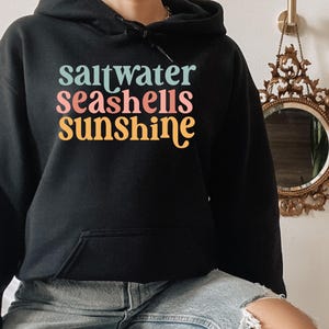 May include: Black hooded sweatshirt with the words "saltwater seashells sunshine" in teal, pink, and yellow. The sweatshirt has a front pocket and a drawstring hood. The text is in a retro font.