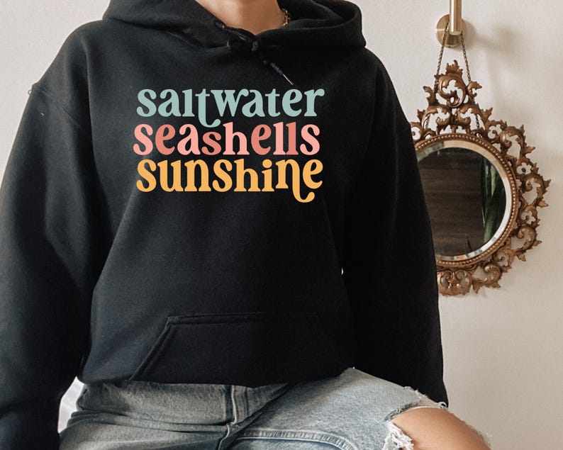 May include: Black hooded sweatshirt with the words "saltwater seashells sunshine" in teal, pink, and yellow. The sweatshirt has a front pocket and a drawstring hood. The text is in a retro font.