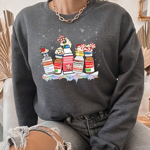 Pu&ograve; includere: Una felpa grigia scura con un design a tema natalizio che presenta cinque bottiglie con diverse etichette, tra cui "Ativan", "Versed", "Peppermint Mocha Propofol", "Midazolam Injection USP" e "Versed". Le bottiglie sono decorate con bastoncini di zucchero, fiocchi di neve e luci natalizie.