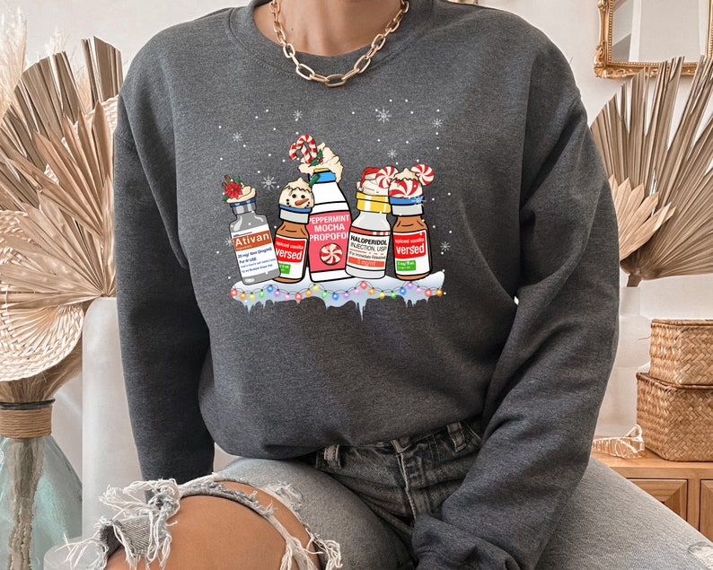 Pu&ograve; includere: Una felpa grigia scura con un design a tema natalizio che presenta cinque bottiglie con diverse etichette, tra cui "Ativan", "Versed", "Peppermint Mocha Propofol", "Midazolam Injection USP" e "Versed". Le bottiglie sono decorate con bastoncini di zucchero, fiocchi di neve e luci natalizie.