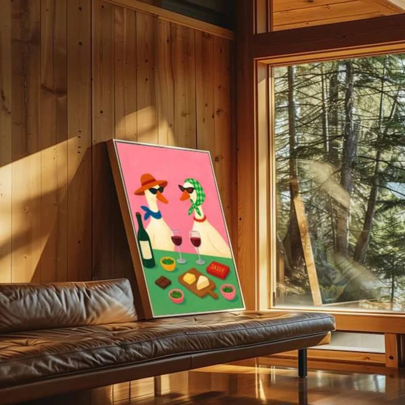 May include: A framed art print featuring two geese wearing sunglasses and hats, enjoying wine and snacks. The artwork has a pink background and a green foreground with the word "Sassy" on a small red sign. The print is leaning against a wooden wall.