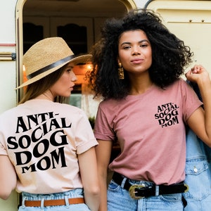 May include: Two women wearing pink and beige t-shirts with the text "Anti-Social Dog Mom" printed in black. The woman on the left is wearing a straw hat and jeans. The woman on the right is wearing jeans and a denim jacket.