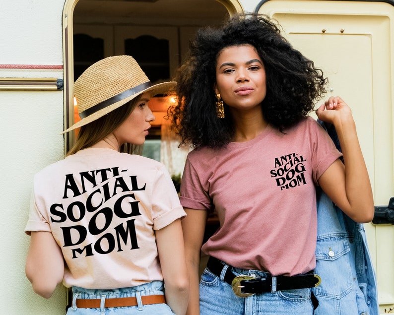 May include: Two women wearing pink and beige t-shirts with the text "Anti-Social Dog Mom" printed in black. The woman on the left is wearing a straw hat and jeans. The woman on the right is wearing jeans and a denim jacket.