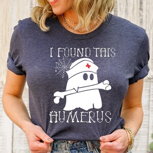 May include: A dark gray t-shirt with a white ghost wearing a nurse's hat and holding a bone. The text "I found this humerus" is printed on the shirt.