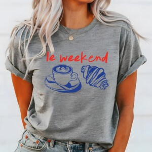 May include: Gray t-shirt with the phrase "le weekend" in red. Below the text is a blue illustration of a coffee cup and a croissant. The t-shirt is a casual style.