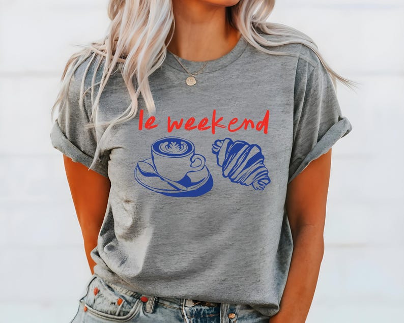 May include: Gray t-shirt with the phrase "le weekend" in red. Below the text is a blue illustration of a coffee cup and a croissant. The t-shirt is a casual style.