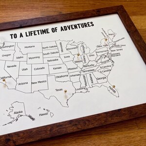 May include: A framed map of the United States with the text TO A LIFETIME OF ADVENTURES printed at the top. The map features black state outlines and labels on a white background, accented with several small gold push pins placed on states.