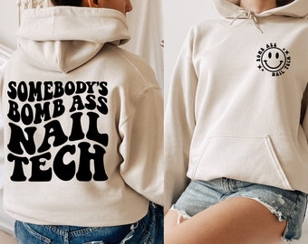 May include: A beige hoodie with the text "Somebody's Bomb Ass Nail Tech" in black on the back and a black smiley face with the text "Bomb Ass Nail Tech" on the front.