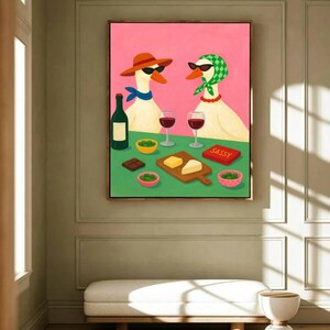 May include: A framed painting depicts two geese wearing sunglasses, hats, and scarves, enjoying wine and snacks. A bottle of wine, cheese, and chocolate are on the table. The word "SASSY" is on a red box. The background is pink and green.