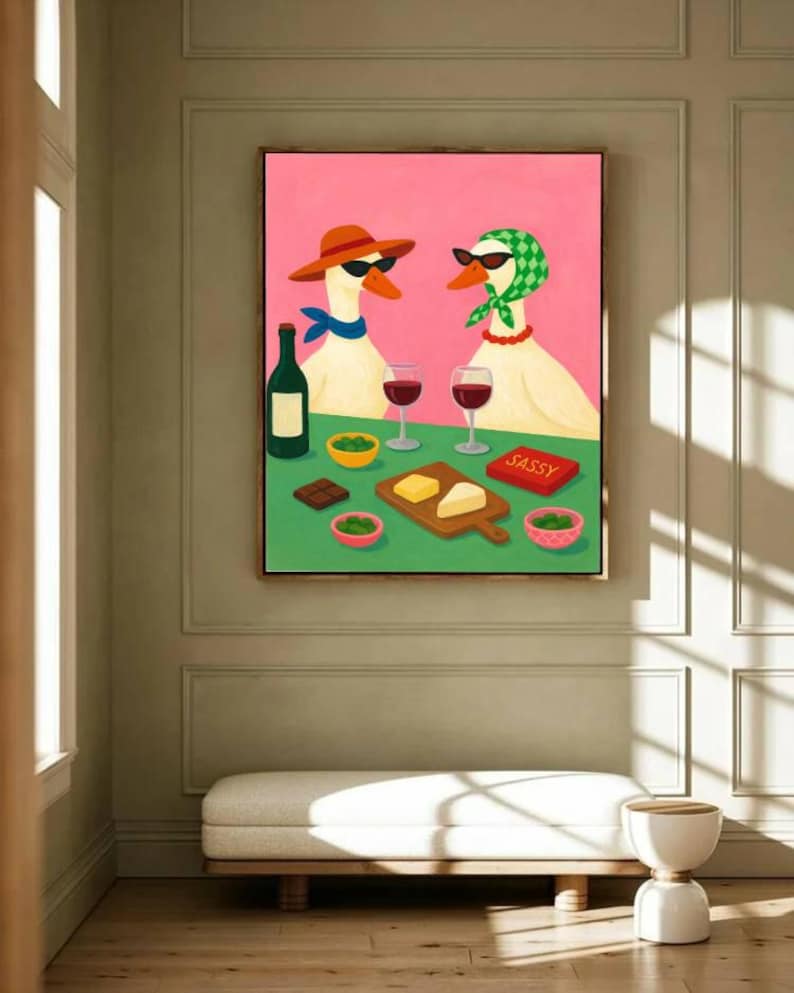 May include: A framed painting depicts two geese wearing sunglasses, hats, and scarves, enjoying wine and snacks. A bottle of wine, cheese, and chocolate are on the table. The word "SASSY" is on a red box. The background is pink and green.