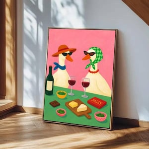 May include: A framed art print featuring two geese wearing sunglasses and accessories, enjoying wine and snacks. The artwork has a pink background and a green table with a bottle of wine, glasses, cheese, and the word "SASSY".