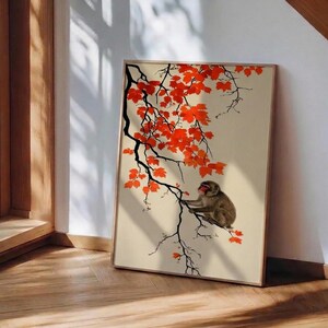 May include: A framed print featuring a Japanese macaque monkey perched on a branch with vibrant red maple leaves. The artwork has a beige background and a wooden frame, creating a serene and natural aesthetic.