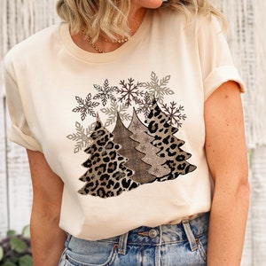 May include: A beige t-shirt with a graphic design of three Christmas trees. The trees are decorated with a leopard print, a brown and white plaid pattern, and a glittery snowflake pattern. The design is surrounded by snowflakes.