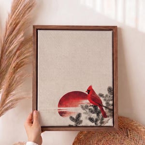 May include: A framed canvas art print featuring a red cardinal perched on a branch, set against a red sunset. The artwork has a neutral background and is encased in a dark brown wooden frame. The style is reminiscent of a watercolor painting.