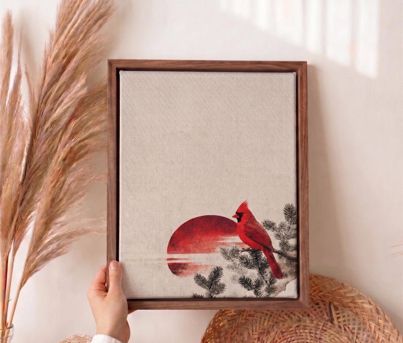 May include: A framed canvas art print featuring a red cardinal perched on a branch, set against a red sunset. The artwork has a neutral background and is encased in a dark brown wooden frame. The style is reminiscent of a watercolor painting.