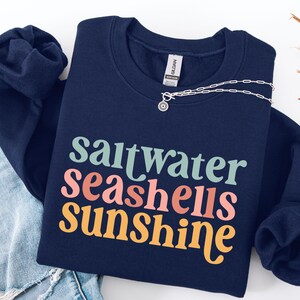 May include: A navy blue sweatshirt with the words "saltwater seashells sunshine" in a colorful, retro font. A silver necklace with a pendant is draped across the sweatshirt. The sweatshirt is folded on top of a pair of blue jeans.
