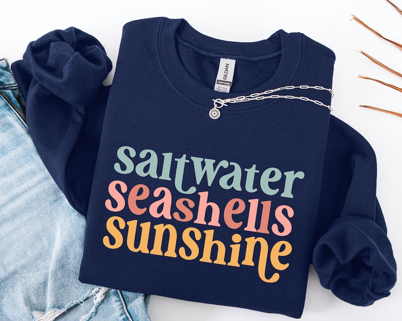 May include: A navy blue sweatshirt with the words "saltwater seashells sunshine" in a colorful, retro font. A silver necklace with a pendant is draped across the sweatshirt. The sweatshirt is folded on top of a pair of blue jeans.
