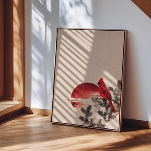 May include: Framed art print featuring a red cardinal perched on a branch, set against a red sun and a neutral background. The artwork is in a wooden frame and leans against a wall, with sunlight casting shadows.