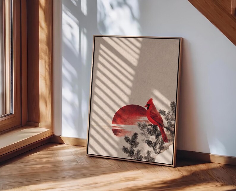 May include: Framed art print featuring a red cardinal perched on a branch, set against a red sun and a neutral background. The artwork is in a wooden frame and leans against a wall, with sunlight casting shadows.