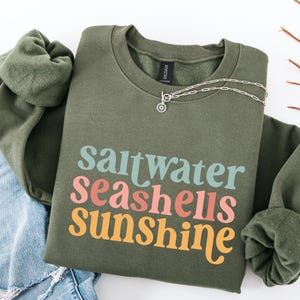May include: An olive green sweatshirt with the words "saltwater seashells sunshine" in teal, coral, and yellow. A silver necklace with a small pendant rests on the shirt. The sweatshirt is paired with blue jeans.