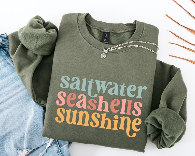 May include: An olive green sweatshirt with the words "saltwater seashells sunshine" in teal, coral, and yellow. A silver necklace with a small pendant rests on the shirt. The sweatshirt is paired with blue jeans.