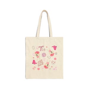 Könnte beinhalten: Eine cremefarbene Tragetasche mit einem Design mit Cartoon-Hunden, die rosa Cowboyhüte und -stiefel tragen. Die Worte "Yee Haw" sind in Pink gedruckt. Weitere rosa und weiße Grafiken sind Schleifen, Sterne und ein Donut.