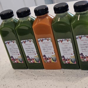 May include: Five bottles of fresh juice with black caps. Four bottles contain green juice, and one contains orange juice. Each bottle has a label with text and fruit illustrations. The bottles are arranged on a light-colored surface.