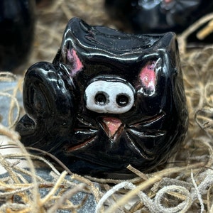 May include: A black ceramic cat figurine with pink ears and a white face. The cat has black eyes and a pink nose.
