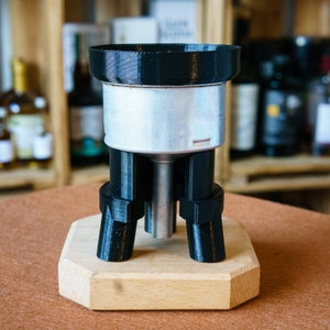 Op de afbeelding: Een zwart 3D-geprint apparaat met een zilveren metalen cilinder in het midden, rustend op een houten basis. Het apparaat heeft drie poten en een trechtervormige bovenkant.