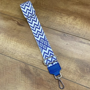 May include: A white and blue patterned strap with a blue leather end and a silver swivel hook. The strap is made of woven fabric and has a geometric design.