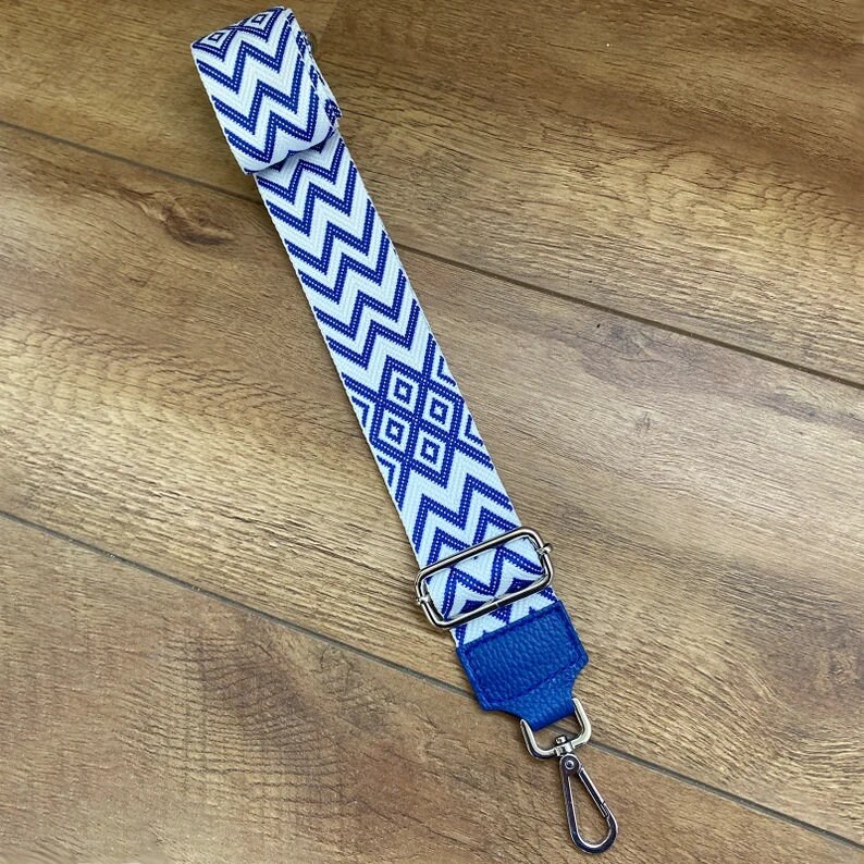 May include: A white and blue patterned strap with a blue leather end and a silver swivel hook. The strap is made of woven fabric and has a geometric design.