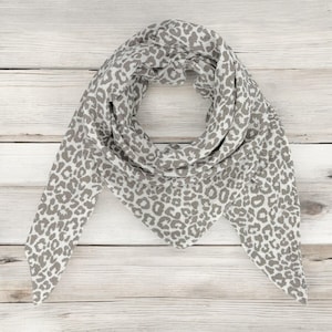 May include: A white and grey scarf with a leopard print pattern. The scarf is folded into a triangular shape and is lying flat on a wooden surface.