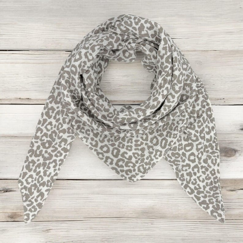 May include: A white and grey scarf with a leopard print pattern. The scarf is folded into a triangular shape and is lying flat on a wooden surface.