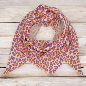May include: A triangular scarf with a leopard print pattern in shades of orange, purple, and beige. The scarf is folded and displayed on a wooden surface.
