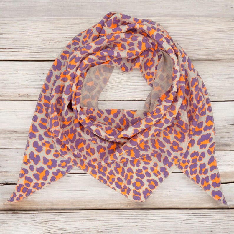 May include: A triangular scarf with a leopard print pattern in shades of orange, purple, and beige. The scarf is folded and displayed on a wooden surface.