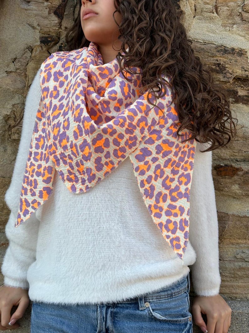 May include: A white fluffy sweater with a purple and orange leopard print scarf. The scarf is tied around the neck and hangs down the front of the sweater.