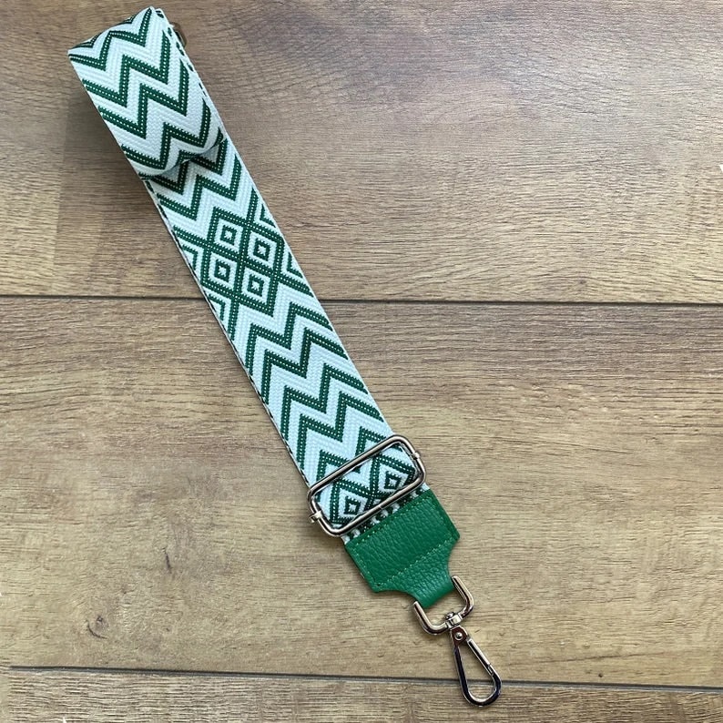 May include: A green and white patterned fabric strap with a green leather end and a silver hook. The strap is designed for a bag or purse.