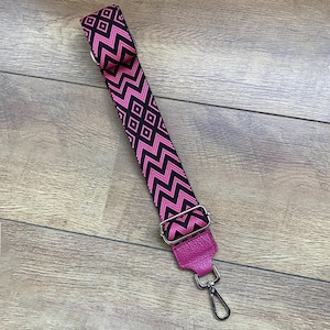 May include: A pink and black patterned adjustable strap with a silver swivel hook. The strap is made of fabric and has a leather end.